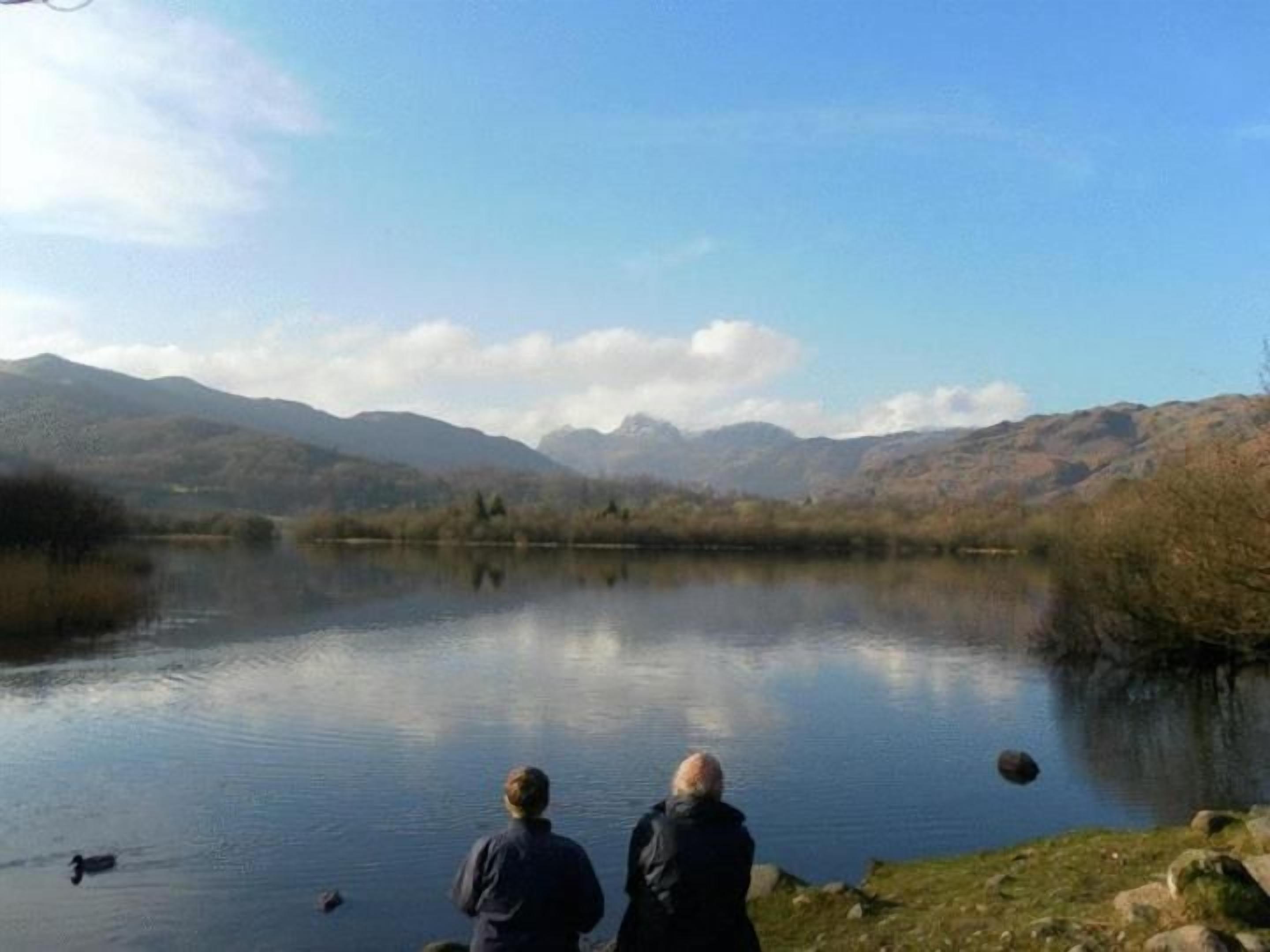 A lake with mountains in the background, a clear sky, and two people standing by the water's edge.