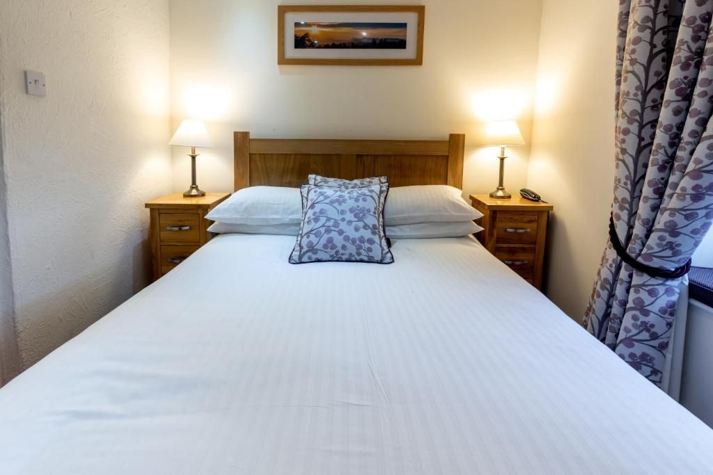 Una cama individual con cabecera de madera, dos mesitas de noche con lámparas, un cuadro enmarcado sobre la cama y una cortina a la derecha.