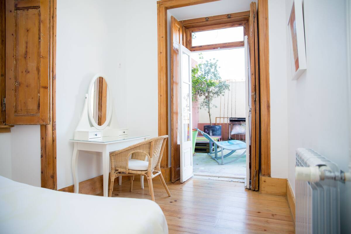 Un dormitorio con suelo de madera, un escritorio blanco con espejo, una silla de mimbre y una cama.