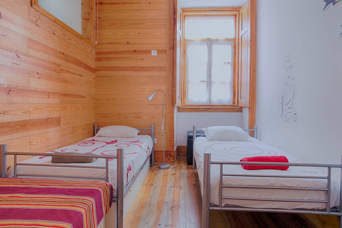 Pequeña habitación con paneles de madera, dos camas individuales, una ventana y un dibujo sencillo en la pared.