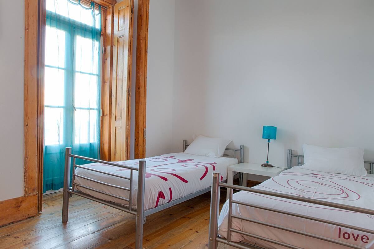 Una habitación con dos literas, piso de madera, una ventana con cortinas y una mesita de noche con lámpara.