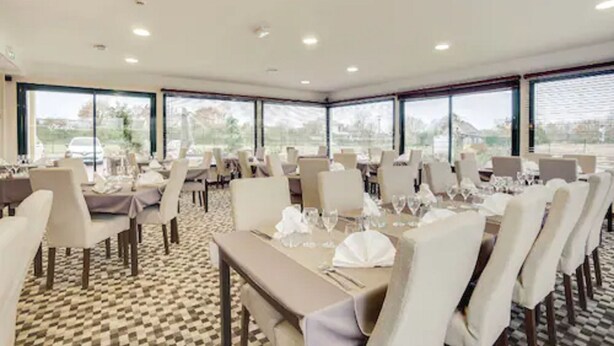 A dining area with multiple tables set for guests, large windows offering a view of the outdoors, and a patterned carpet on the floor. - Brit Hotel La Cote Des Havres (Lessay)