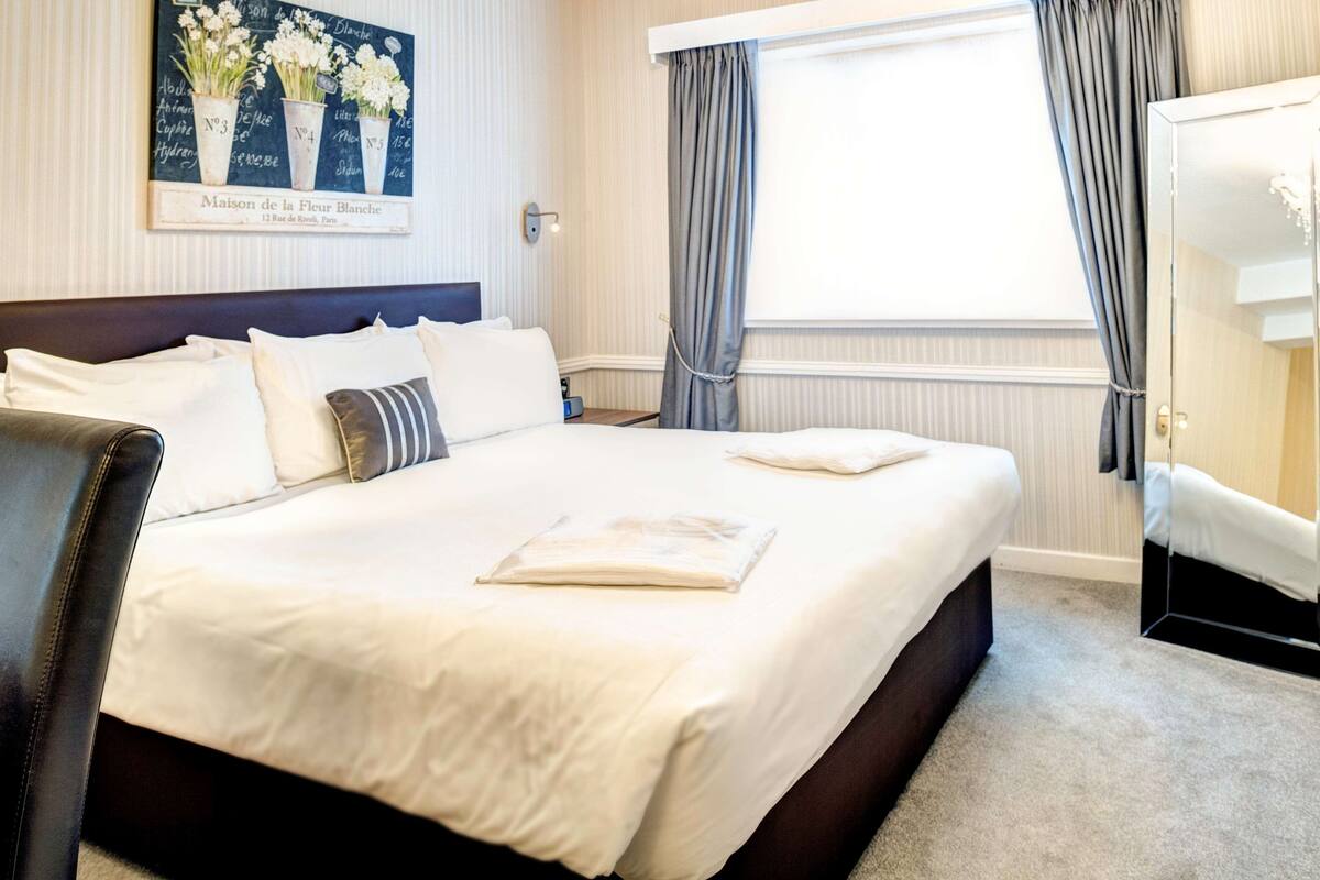 A hotel room with a large bed, a headboard, a bedside table, a mirror, and a window with curtains.