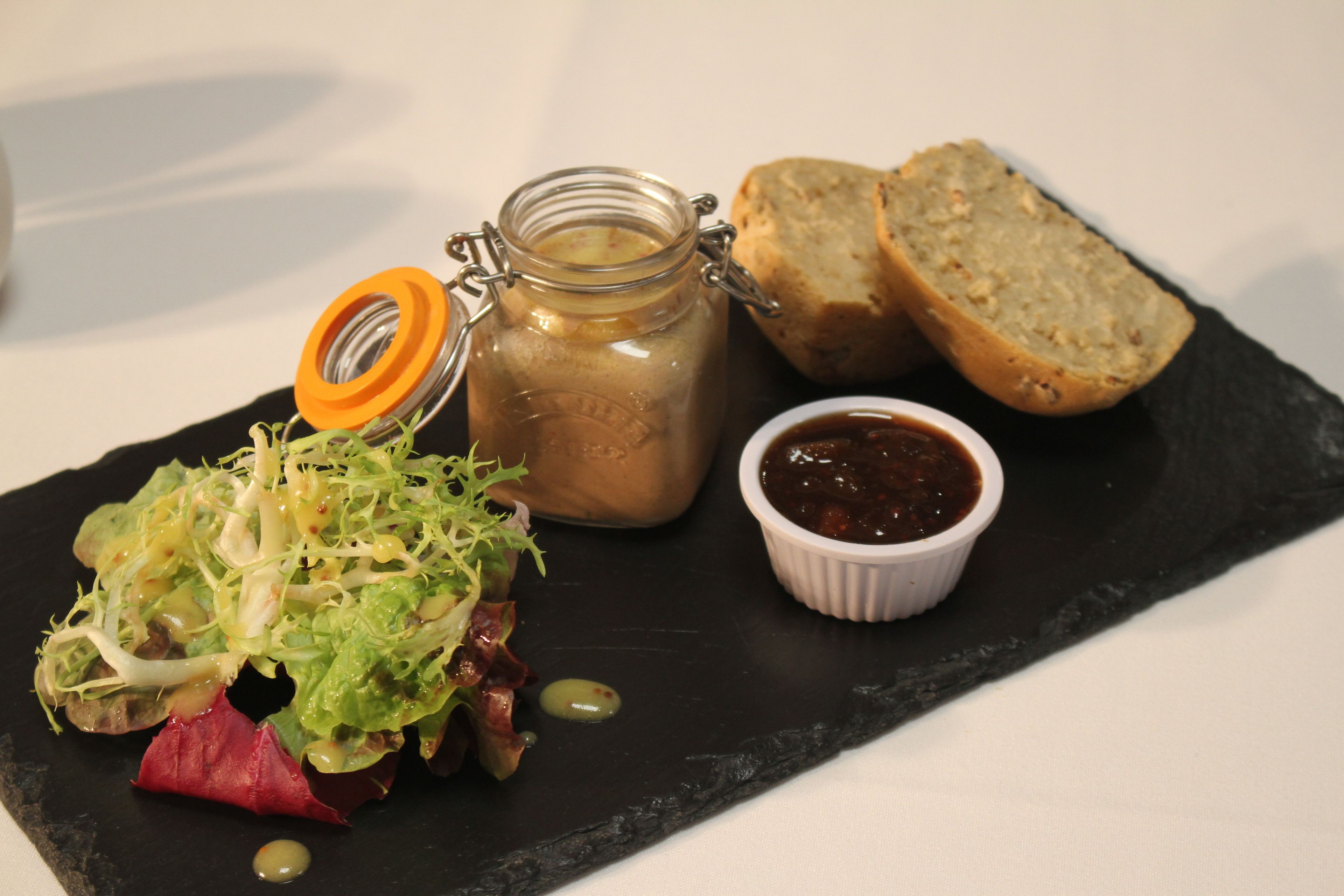 Eine schwarze Schieferplatte mit Salat, einem Senfglas, Brotscheiben und einer kleinen Schale Chutney.