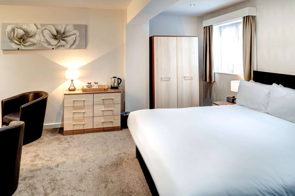 A hotel room with a large bed, a dresser with drawers, a chair, a desk with a lamp, a window with curtains, and a wall-mounted artwork.