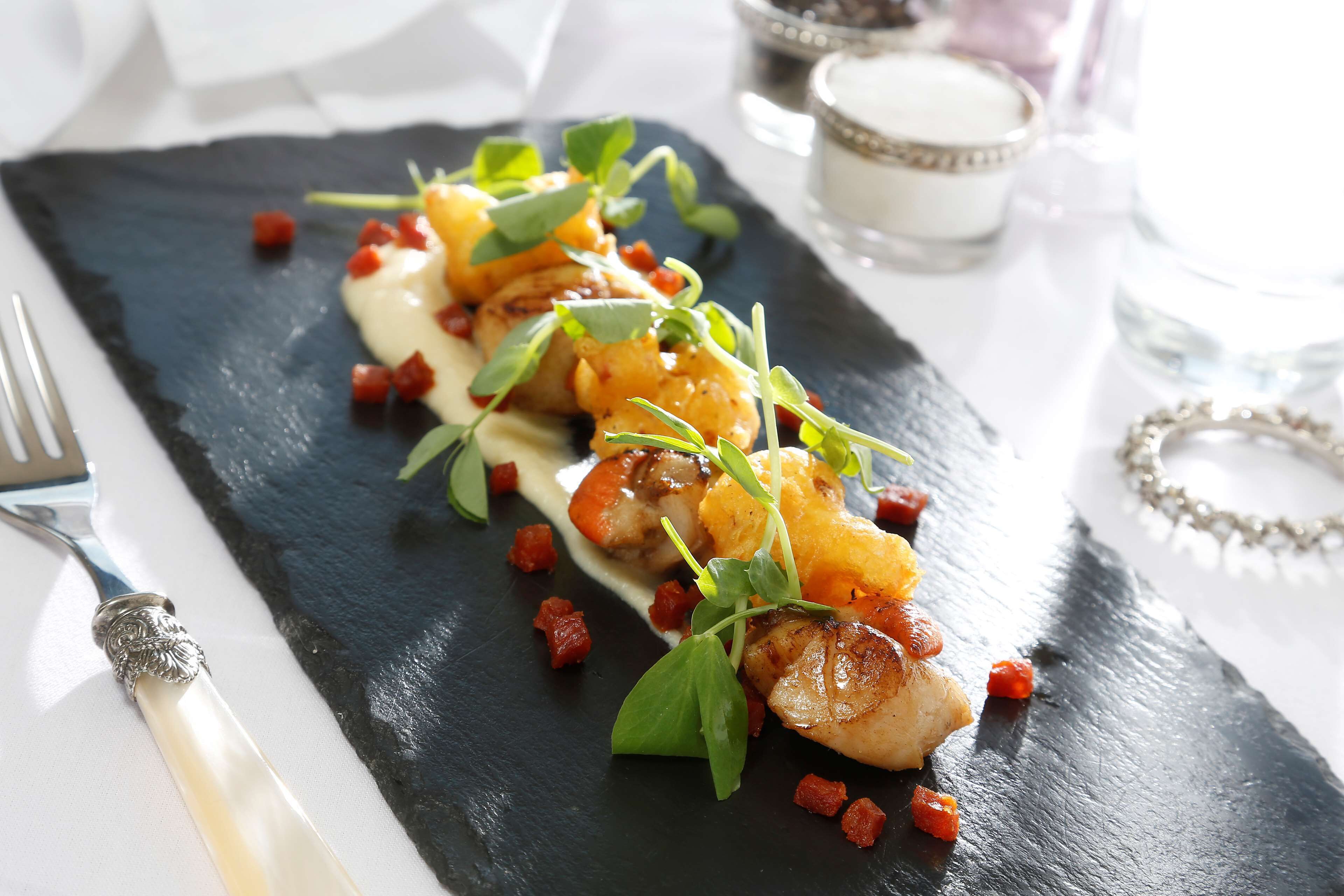 A black slate plate with a dish of seared scallops, garnished with microgreens and diced tomatoes, accompanied by a white sauce.
