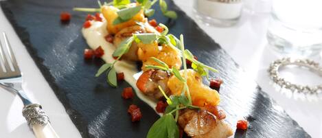 A black slate plate with a dish of seared scallops, garnished with microgreens and diced tomatoes, accompanied by a white sauce.