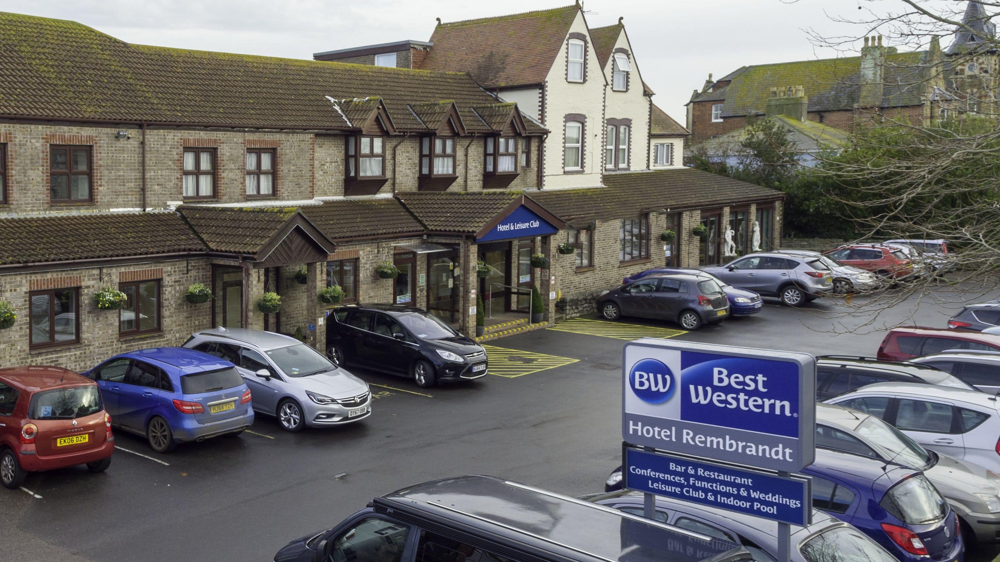 Un hotel de estilo arquitectónico tradicional, con una entrada destacada que muestra un letrero azul. Varias coches están estacionados frente al hotel, que parece tener una zona de estacionamiento.