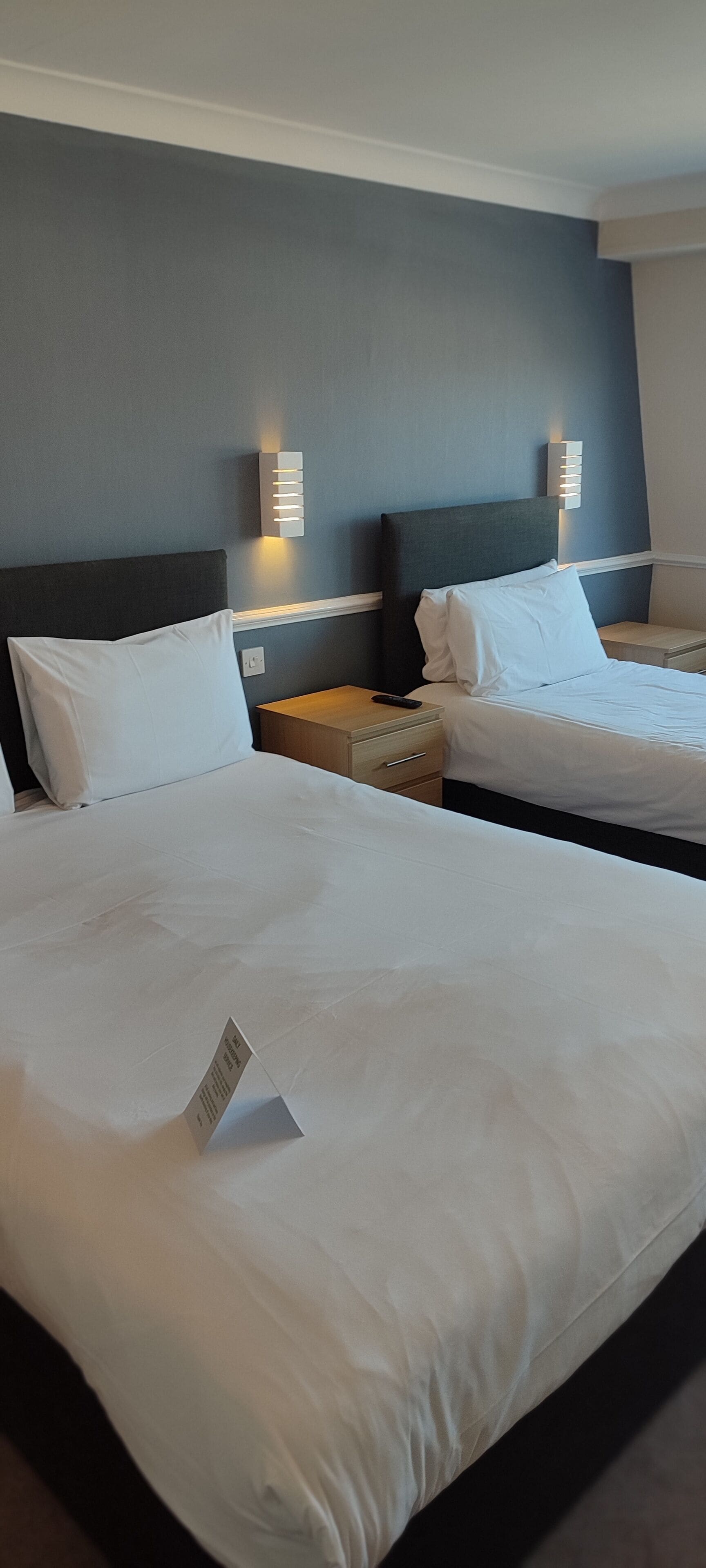 Una habitación de hotel con dos camas, cada una con ropa de cama blanca y cabeceros oscuros. Hay lámparas de pared a ambos lados de las camas.