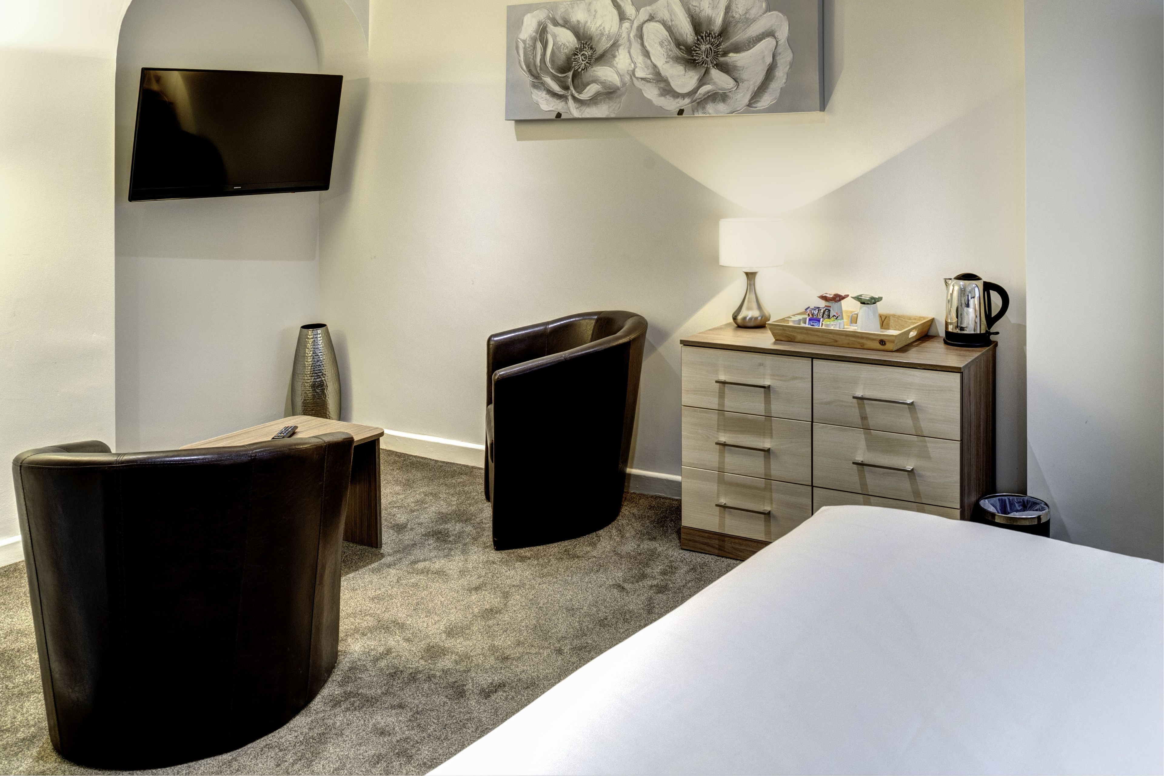 Una camera d'albergo con un letto, una TV a muro, una scrivania con una lampada, un cestino e un quadro decorativo con dei fiori.