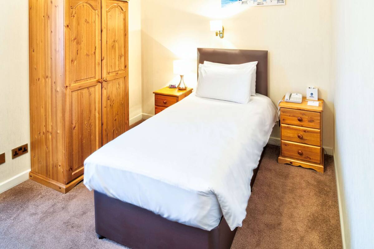A neatly made bed with a white comforter and pillows, a wooden wardrobe, a bedside table with a lamp, and a small drawer unit.