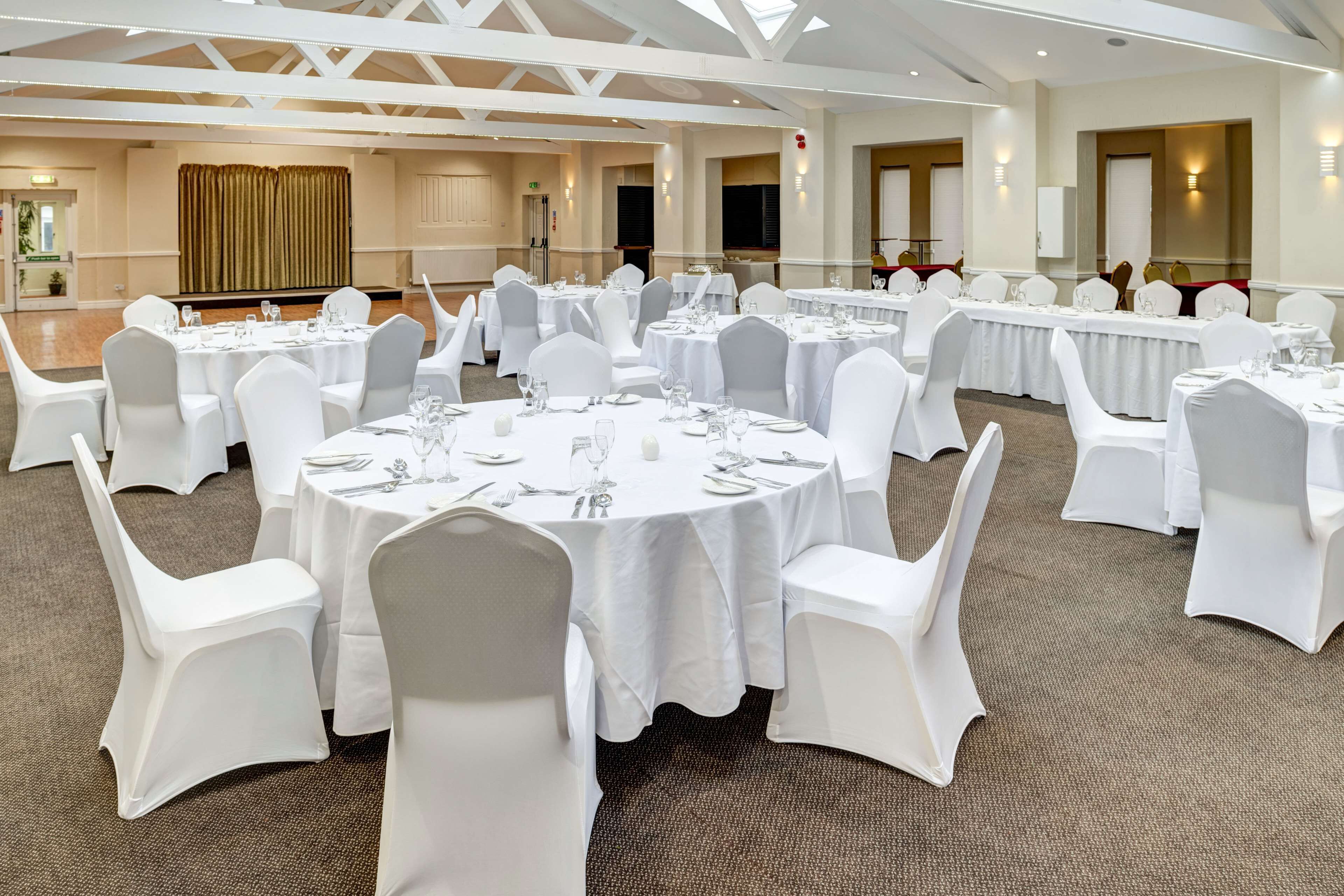 A banquet hall with round tables set for dining, white chairs, and a high ceiling with exposed beams.