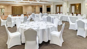 A banquet hall with round tables set for dining, white chairs, and a high ceiling with exposed beams.