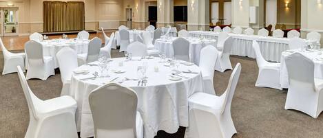 A banquet hall with round tables set for dining, white chairs, and a high ceiling with exposed beams.