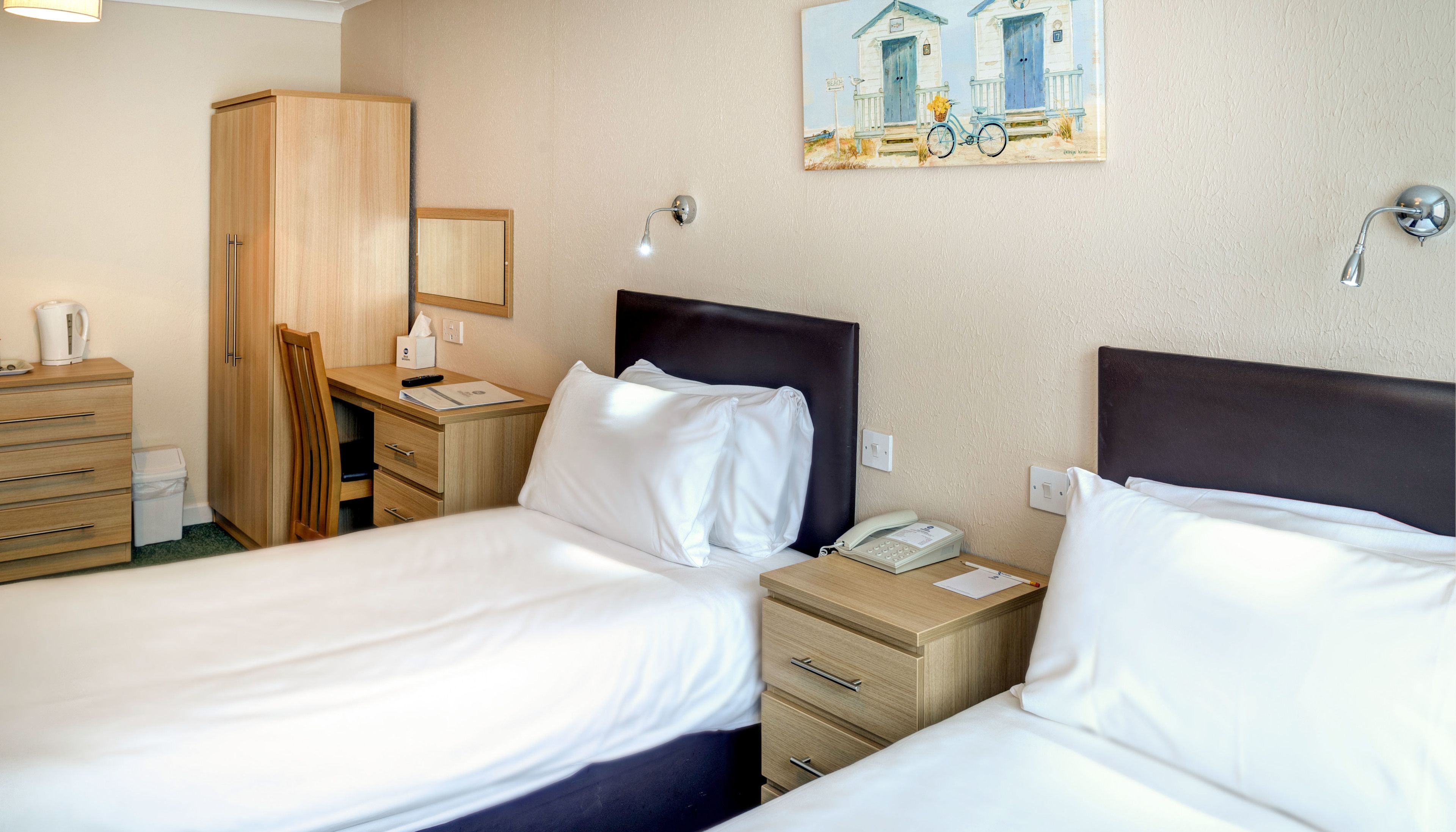 A hotel room with two beds, a desk, a wardrobe, and a painting on the wall.