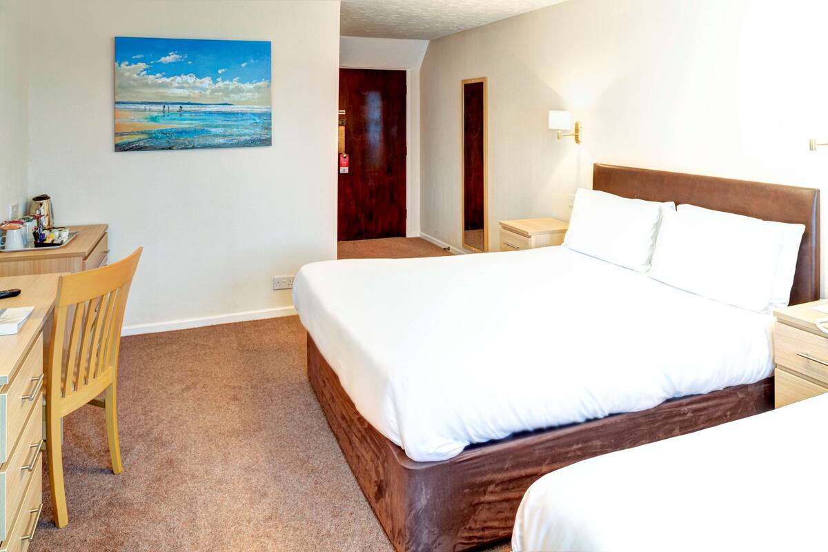 A hotel room with a large bed, a desk with a chair, a painting of a beach, and a door leading to another room.