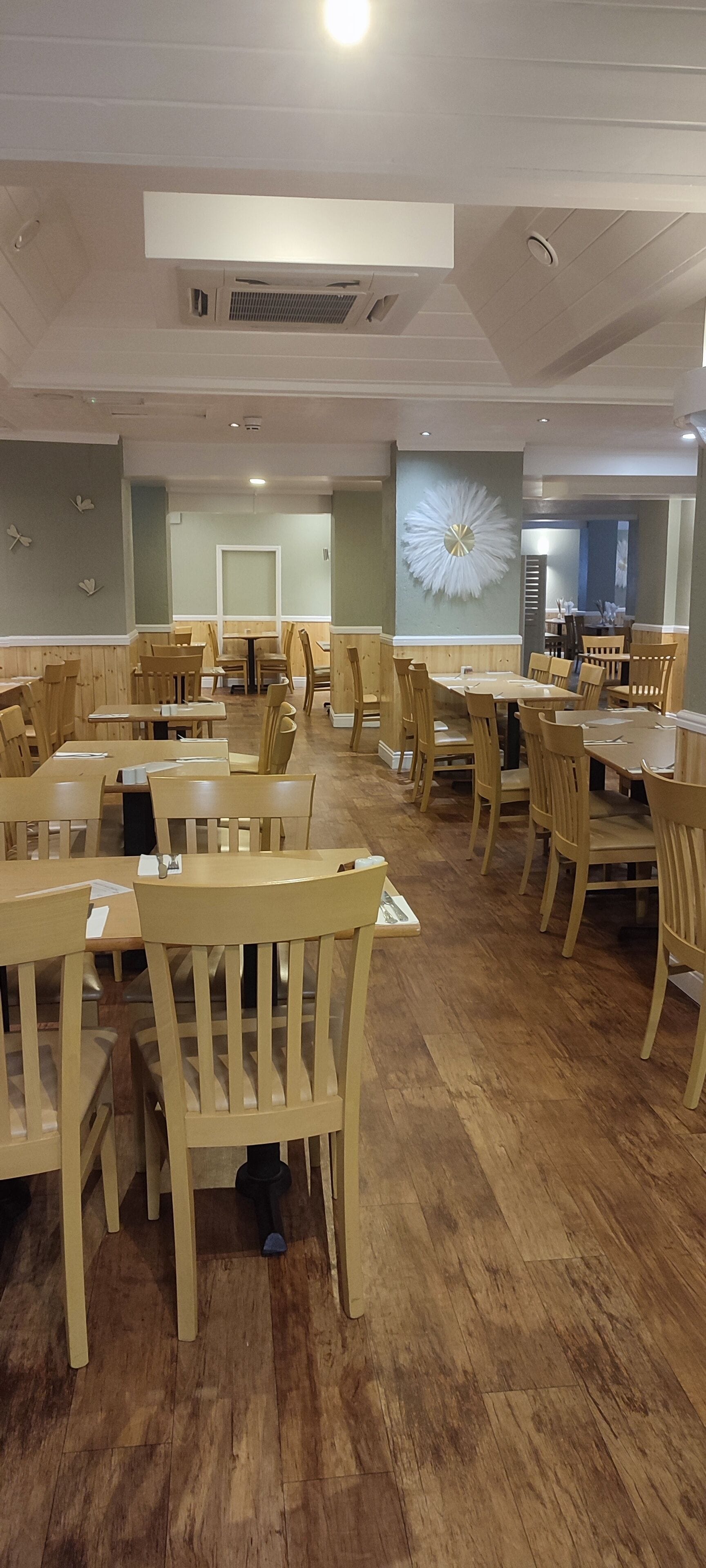 A spacious dining area with wooden tables and chairs, a decorative wall piece, and a ceiling vent.