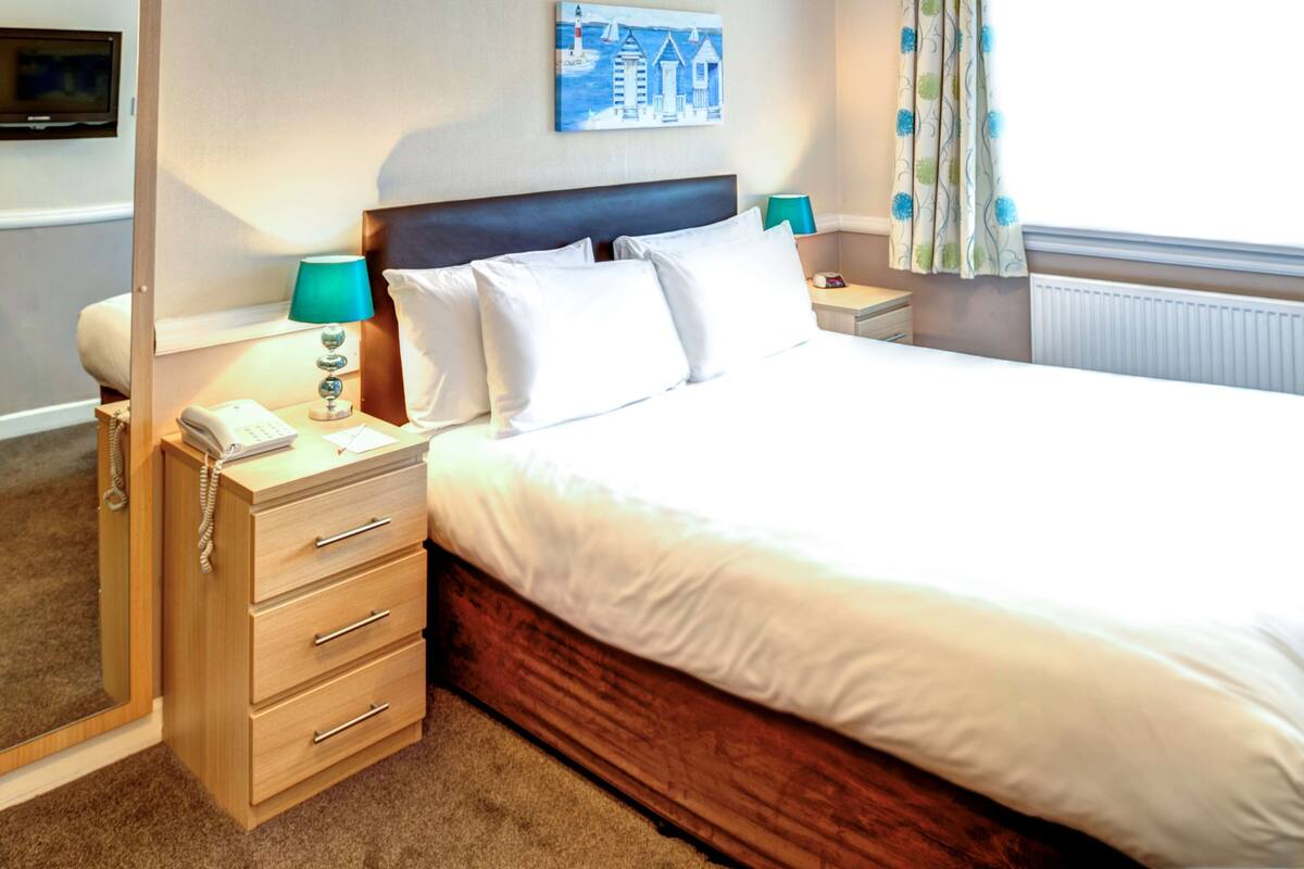 A neatly made bed with white linens, a wooden headboard, and a bedside table with a lamp and a telephone.