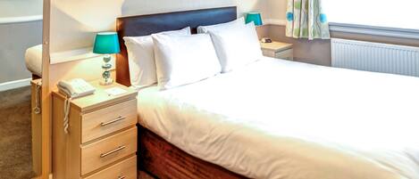 A neatly made bed with white linens, a wooden headboard, and a bedside table with a lamp and a telephone.