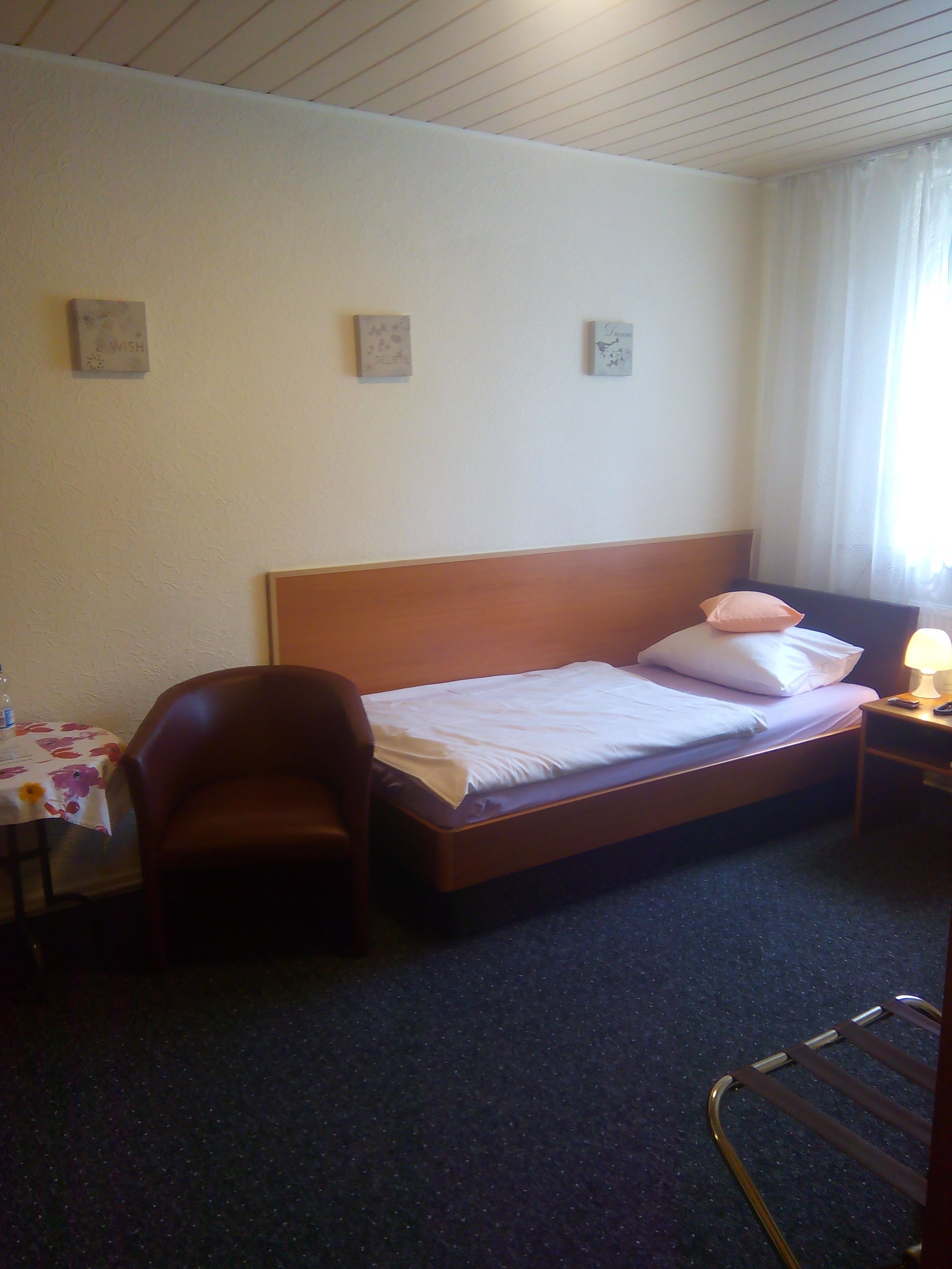 A hotel room with a single bed, a chair, a table with a floral tablecloth, and a nightstand with a lamp.