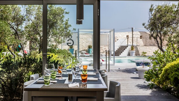 An outdoor dining area with a pool view, tables set with glasses and napkins, and a modern architectural design. - Hotel Genovese (Bonifacio)