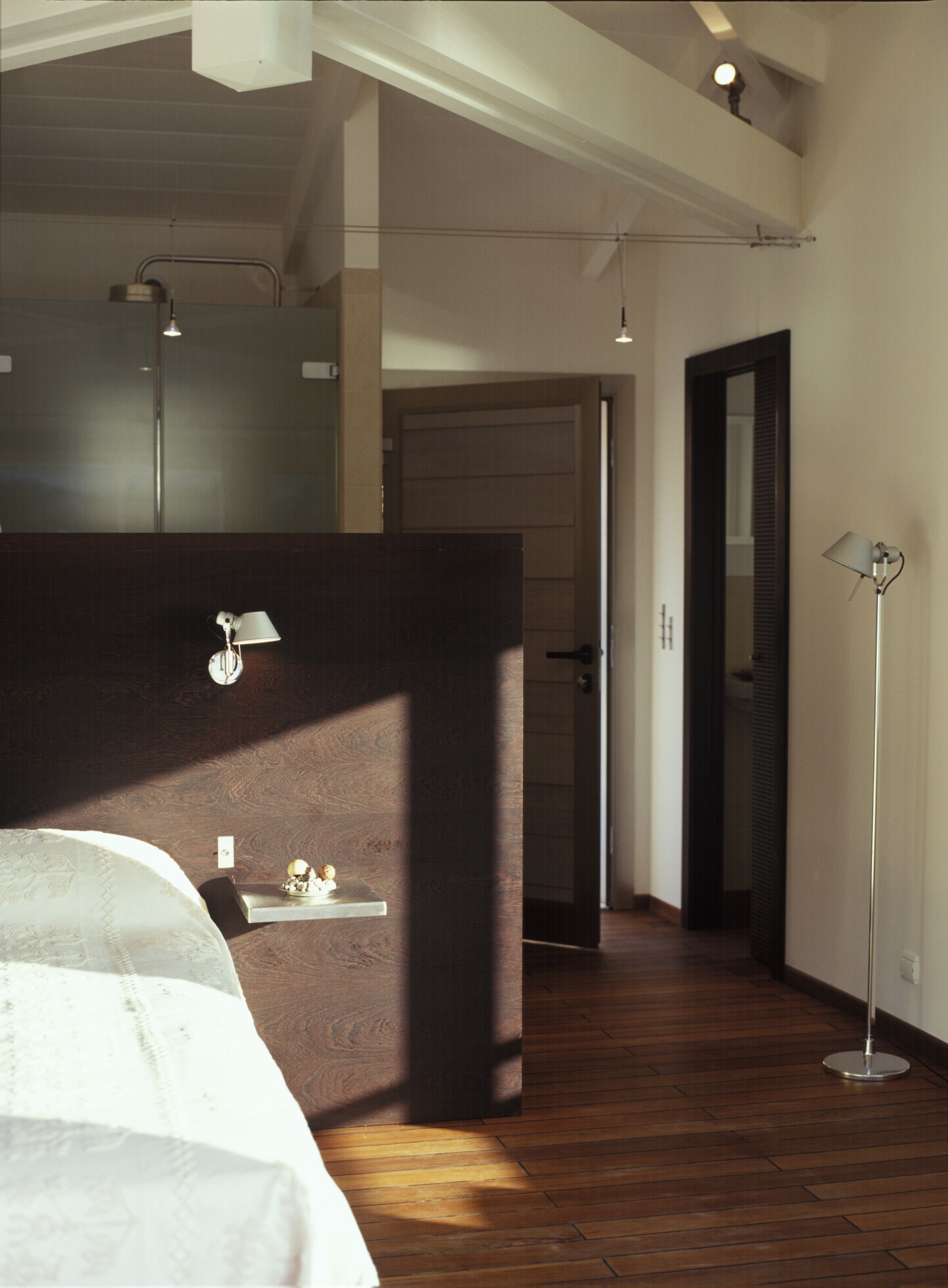 Une chambre avec un grand lit, une table de chevet, un bureau et une lampe sur pied. Une porte donne accès à une autre pièce.