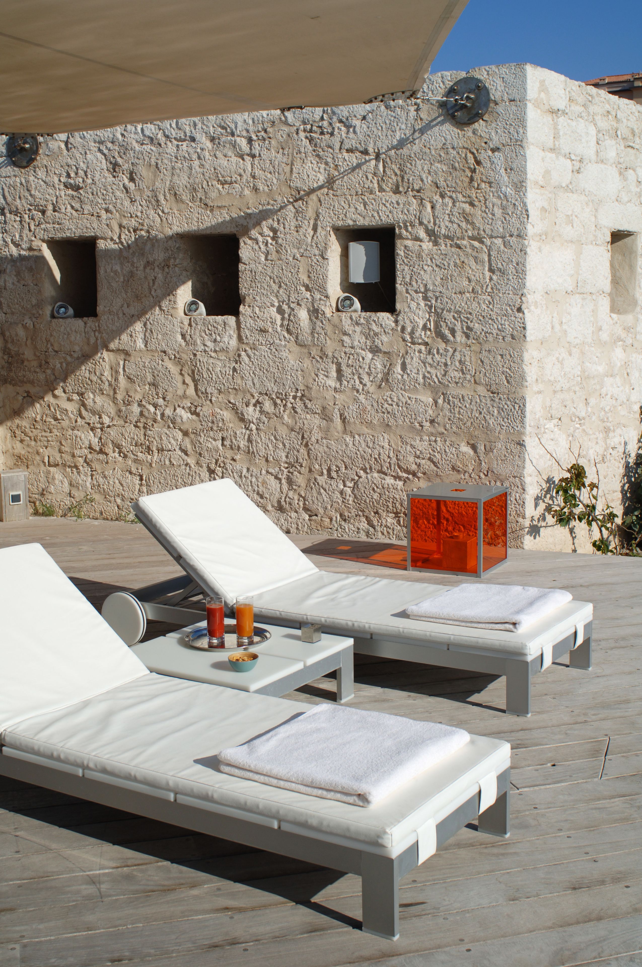 Two white sun loungers with towels and a glass of orange juice on a wooden deck, against a stone wall with small windows.
