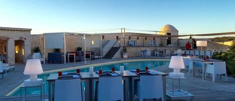 A poolside area with tables and chairs, a stone wall, and a staircase leading to an upper level.