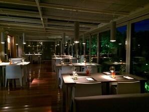 Um restaurante com mesas e cadeiras compridas, janelas grandes e teto alto com vigas aparentes.