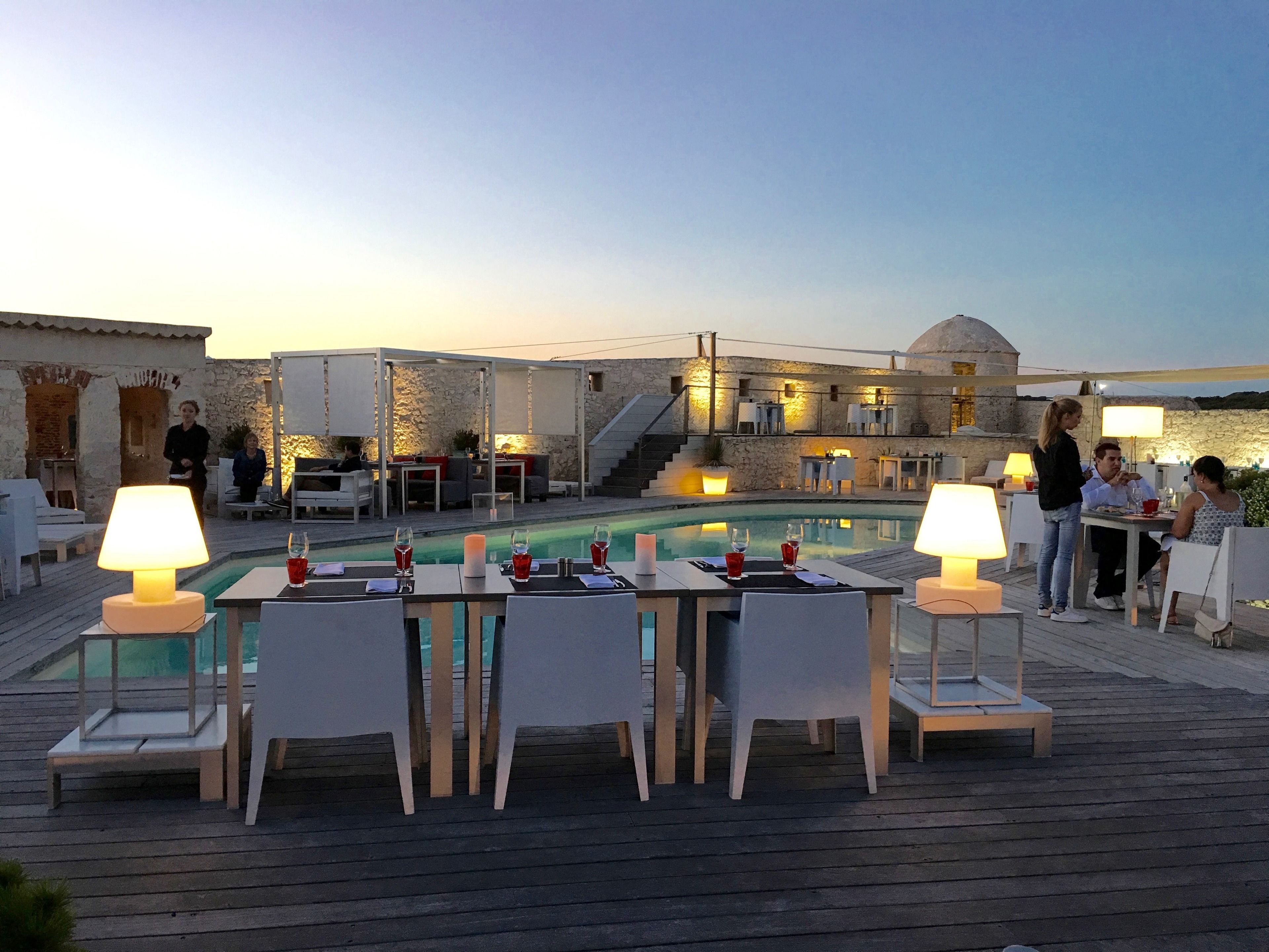 An outdoor dining area with tables and chairs, a pool, and a stone building in the background.