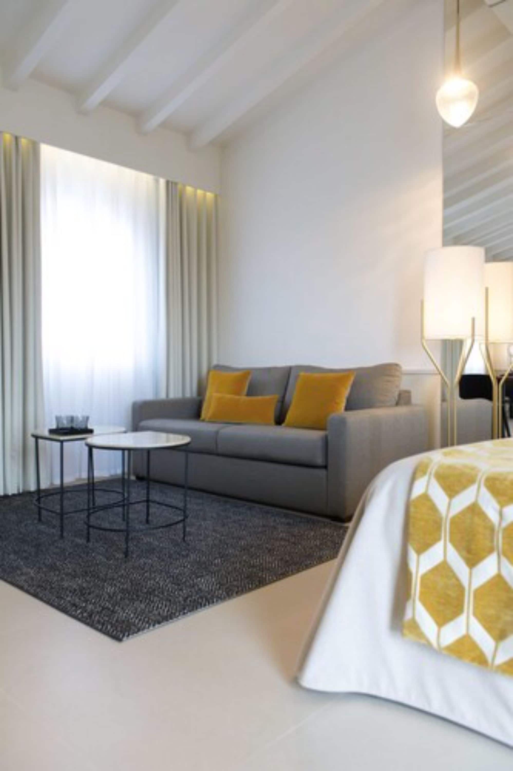 A modern hotel room with a grey sofa, a small round table, and a bed with a patterned bedspread.