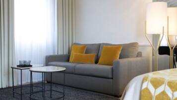 A modern hotel room with a grey sofa, a small round table, and a bed with a patterned bedspread.