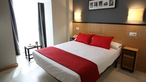 Habitación de hotel con una cama grande, ropa de cama roja y blanca, una mesita de noche con una lámpara y una foto enmarcada en la pared.