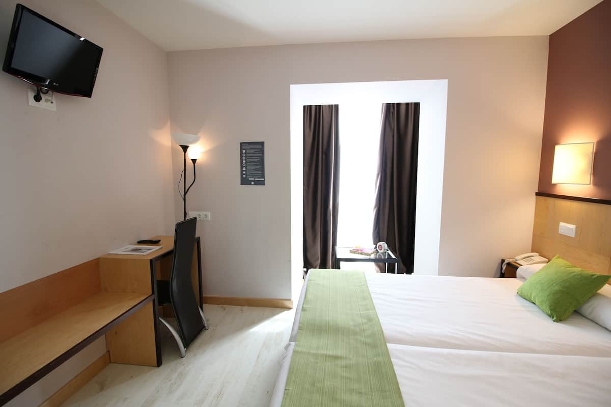 A hotel room with a bed, a desk, a TV, a lamp, a phone, and a window with curtains.