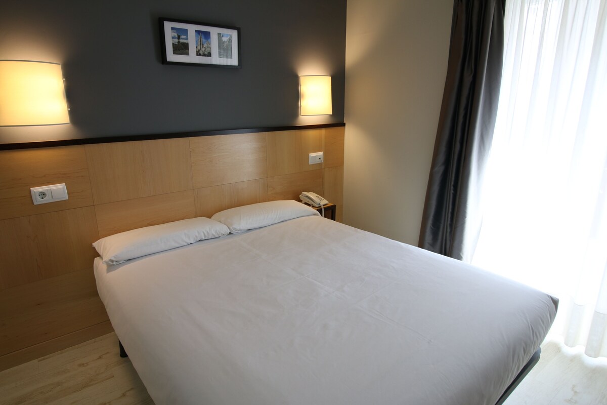A neatly made bed with white linens, a bedside telephone, and two wall-mounted light fixtures.