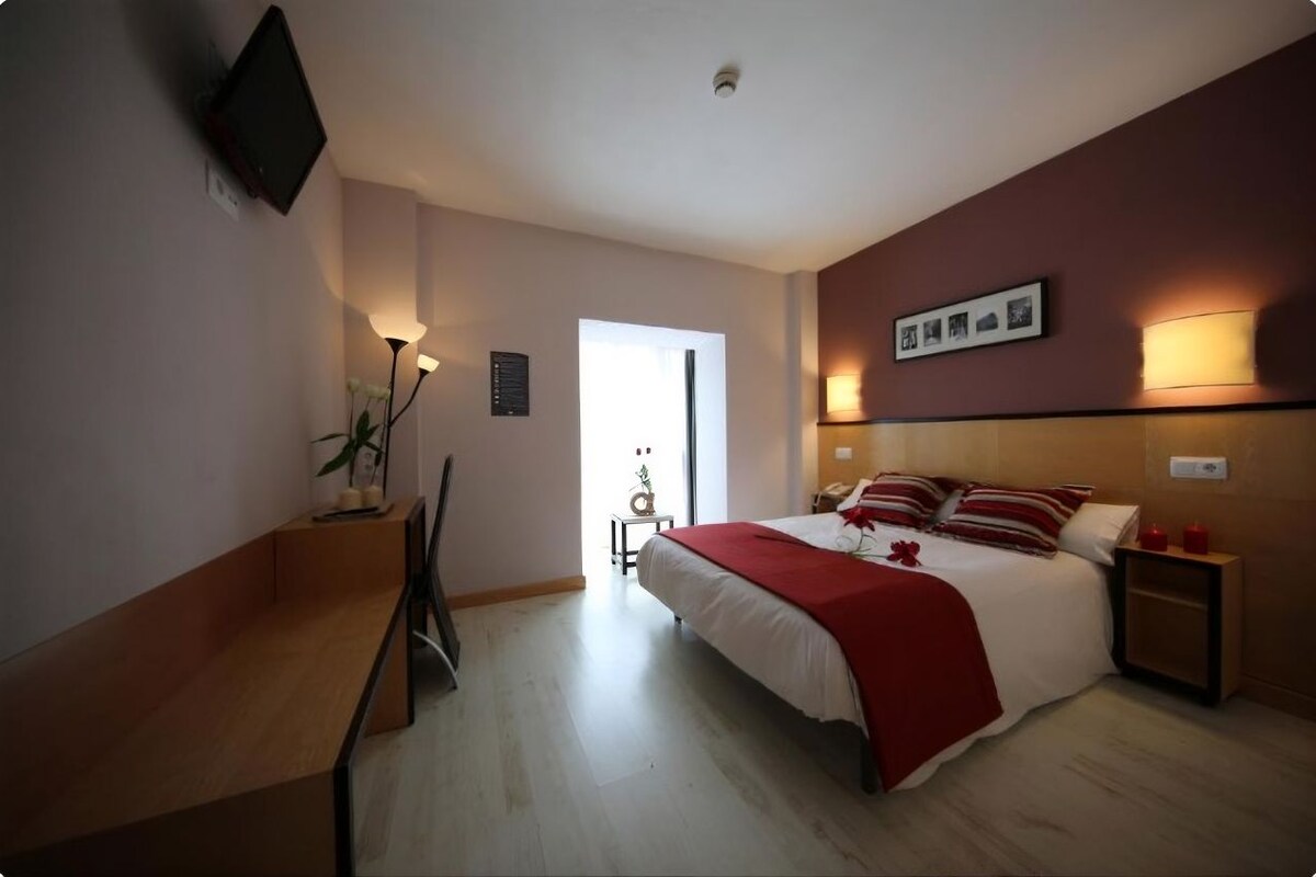 A hotel room with a bed, bedside tables, a desk, a chair, a TV mounted on the wall, and a door leading to another room.