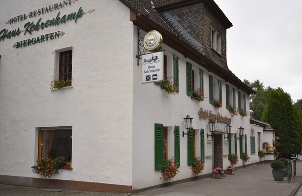 Un bâtiment traditionnel, de couleur blanche, avec des volets verts, une enseigne indiquant « Hôtel-restaurant Haus Kehrenkamp, Biergarten », et des jardinières aux fenêtres.