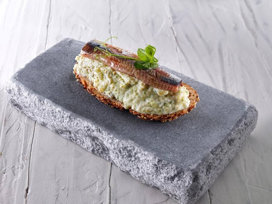 A slice of bread with a creamy spread and a piece of fish on top, placed on a gray stone slab.