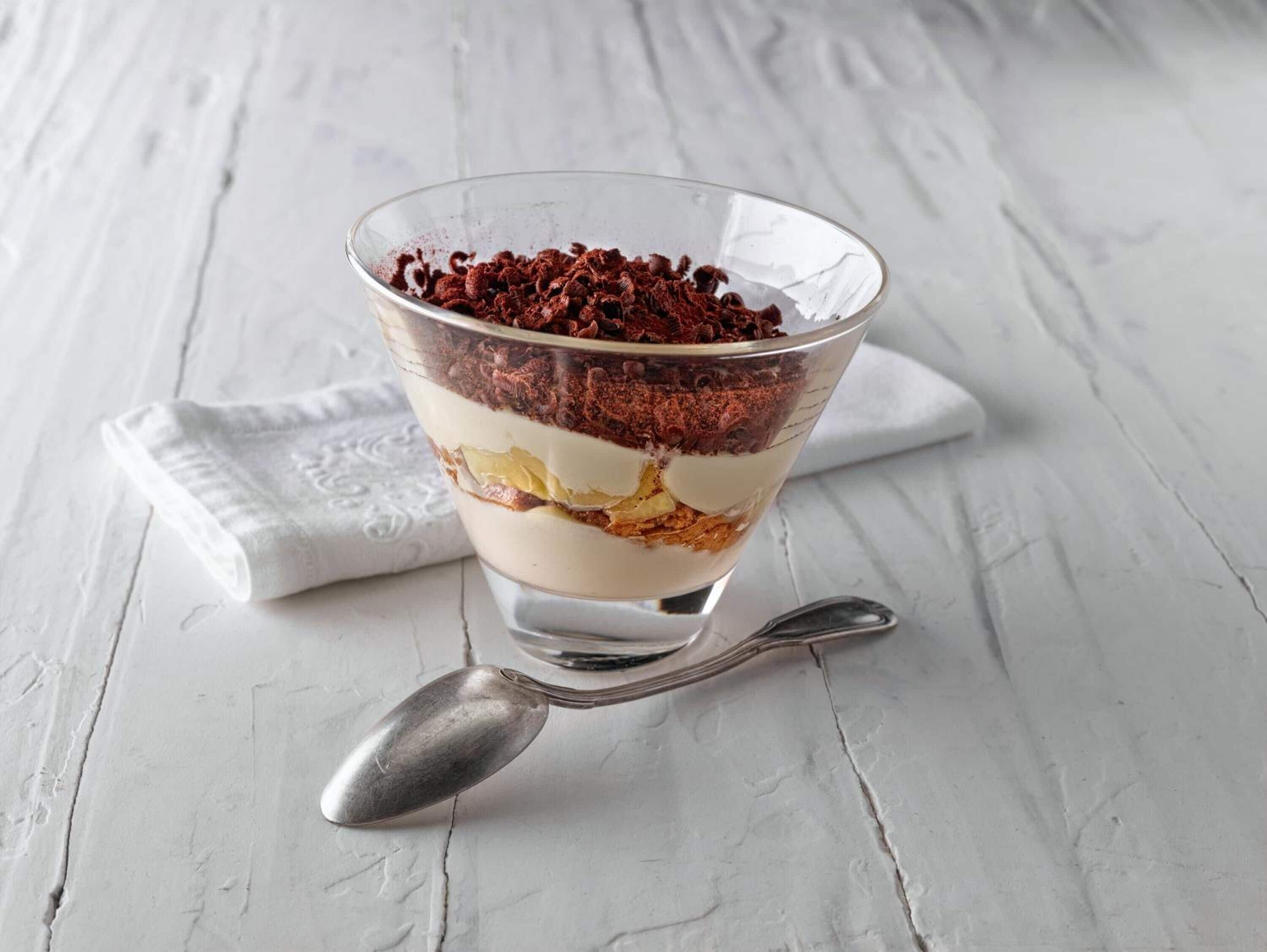 A layered dessert in a clear glass bowl on a white textured surface with a spoon beside it.