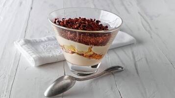 A layered dessert in a clear glass bowl on a white textured surface with a spoon beside it.