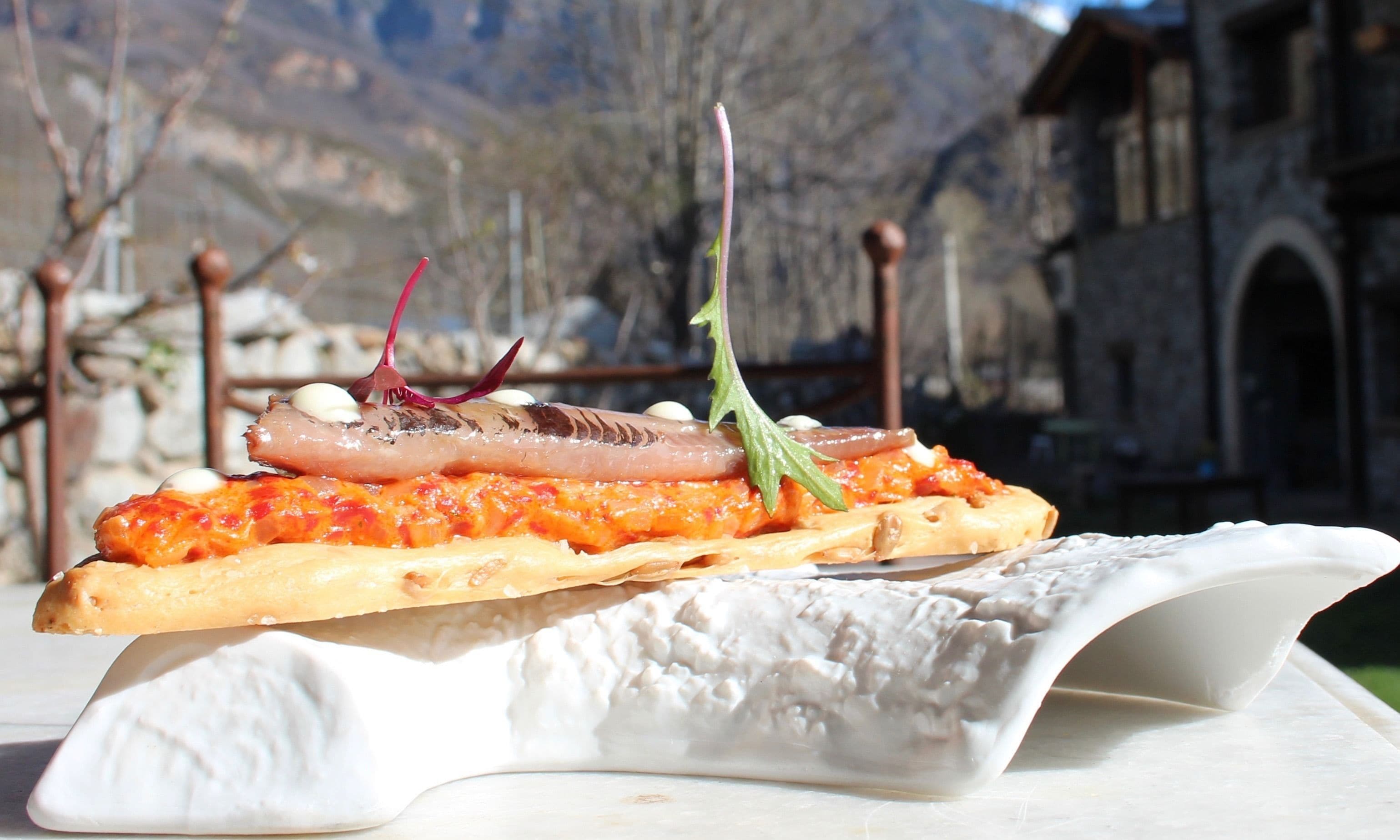 Un plato con filete de pescado, salsa de tomate y decoración sobre un pan baguette, con un paisaje montañoso de fondo.