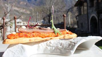 Un plato con filete de pescado, salsa de tomate y decoración sobre un pan baguette, con un paisaje montañoso de fondo.