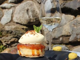 Un postre con una capa caramelizada y una ramita de menta, acompañado de una copa de vino blanco, sobre un fondo de pared de piedra.