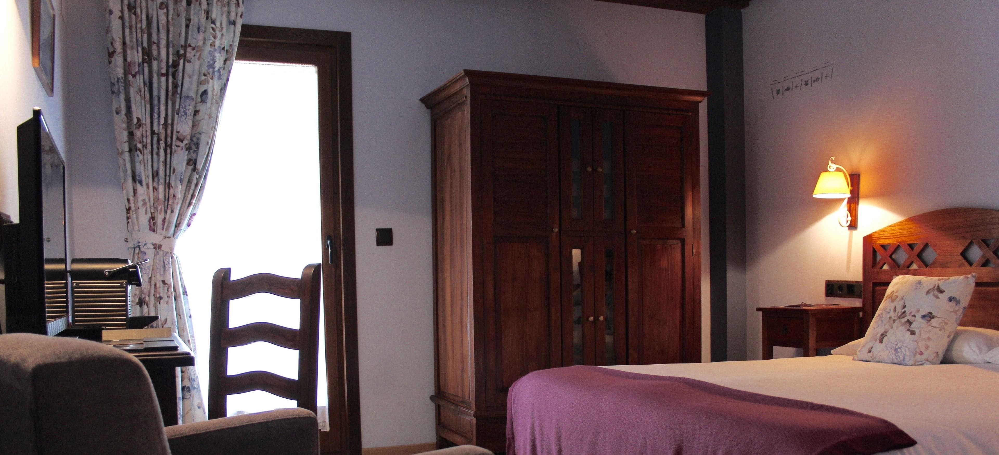 A bedroom with a bed, a wooden wardrobe, a desk with a chair, and a lamp.