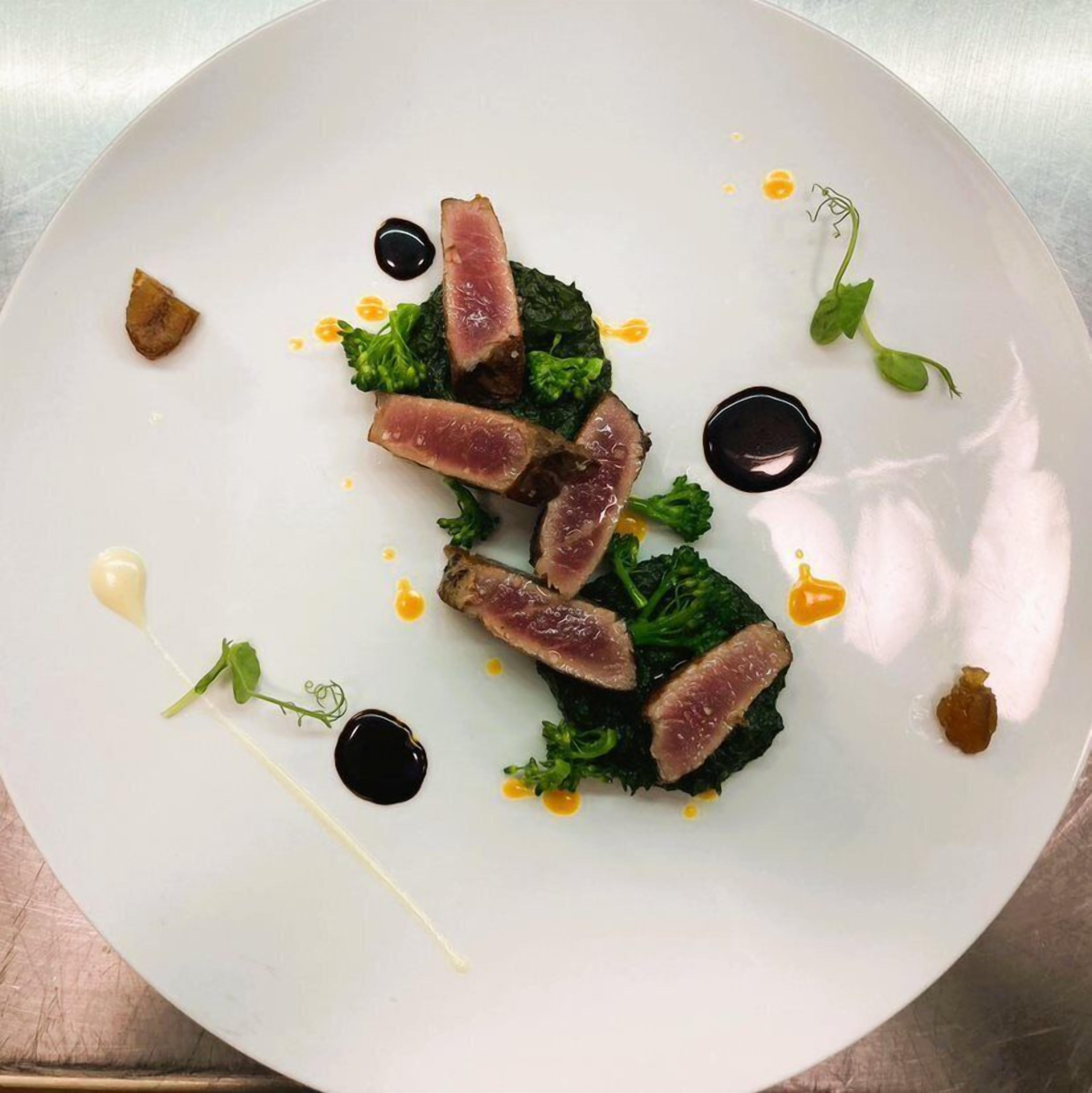 A white plate with a dish of sliced meat, greens, and sauce.