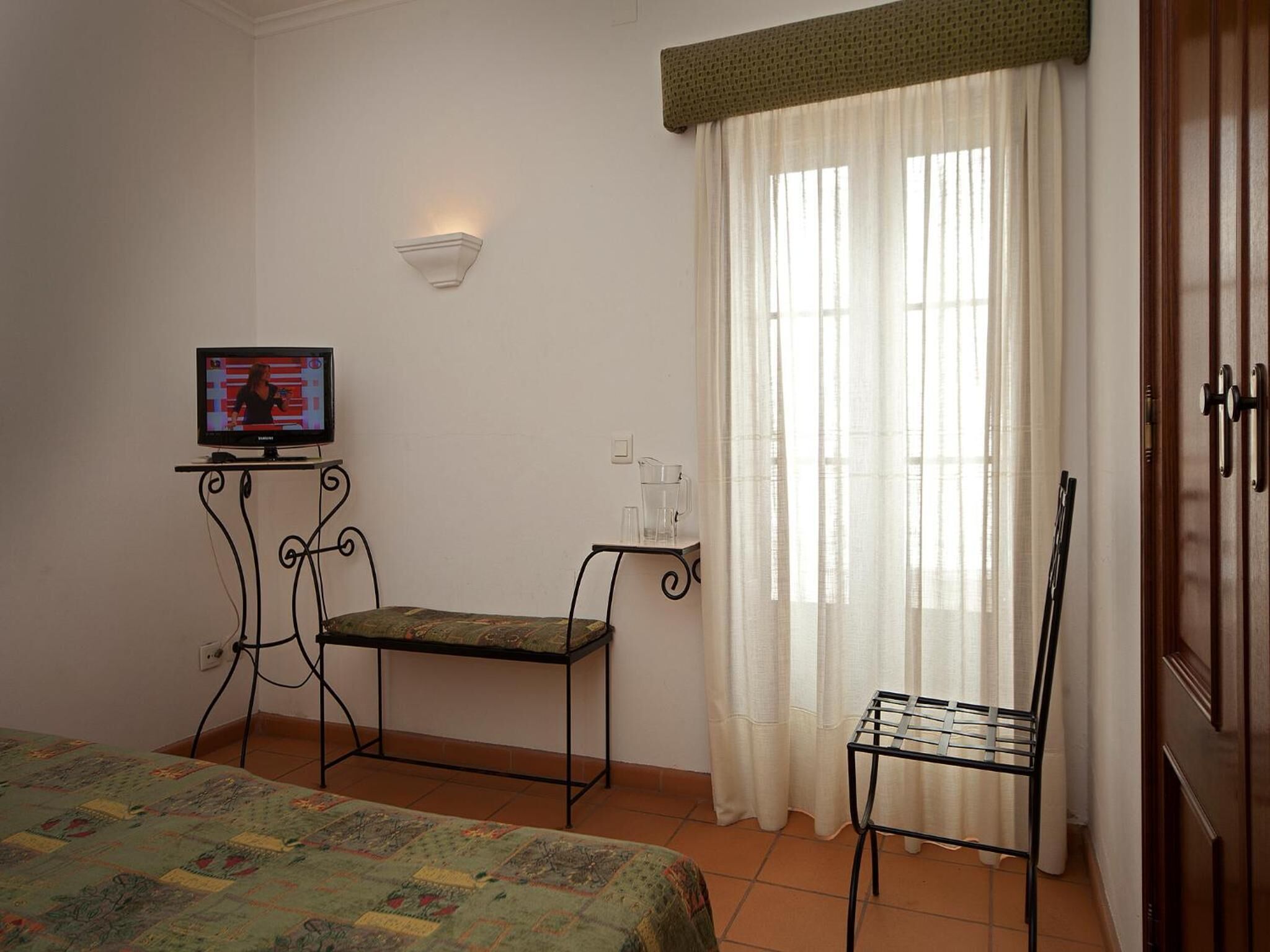 Una habitación pequeña con una cama, un televisor sobre un mueble bajo, una mesa con una silla y una ventana con cortinas.