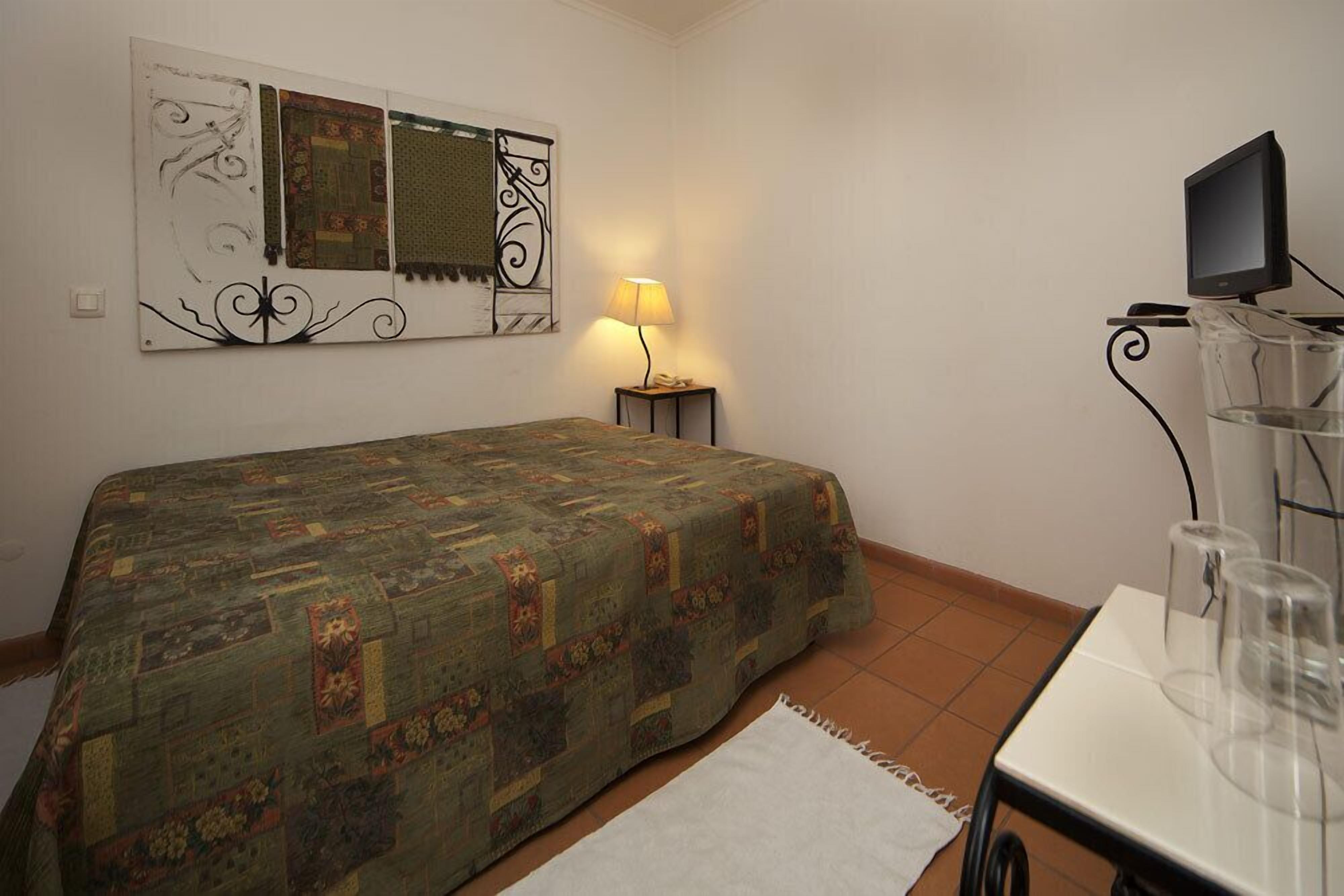 Un dormitorio pequeño y ordenado con cama, escritorio con computadora, lámpara y televisor sobre un mueble.