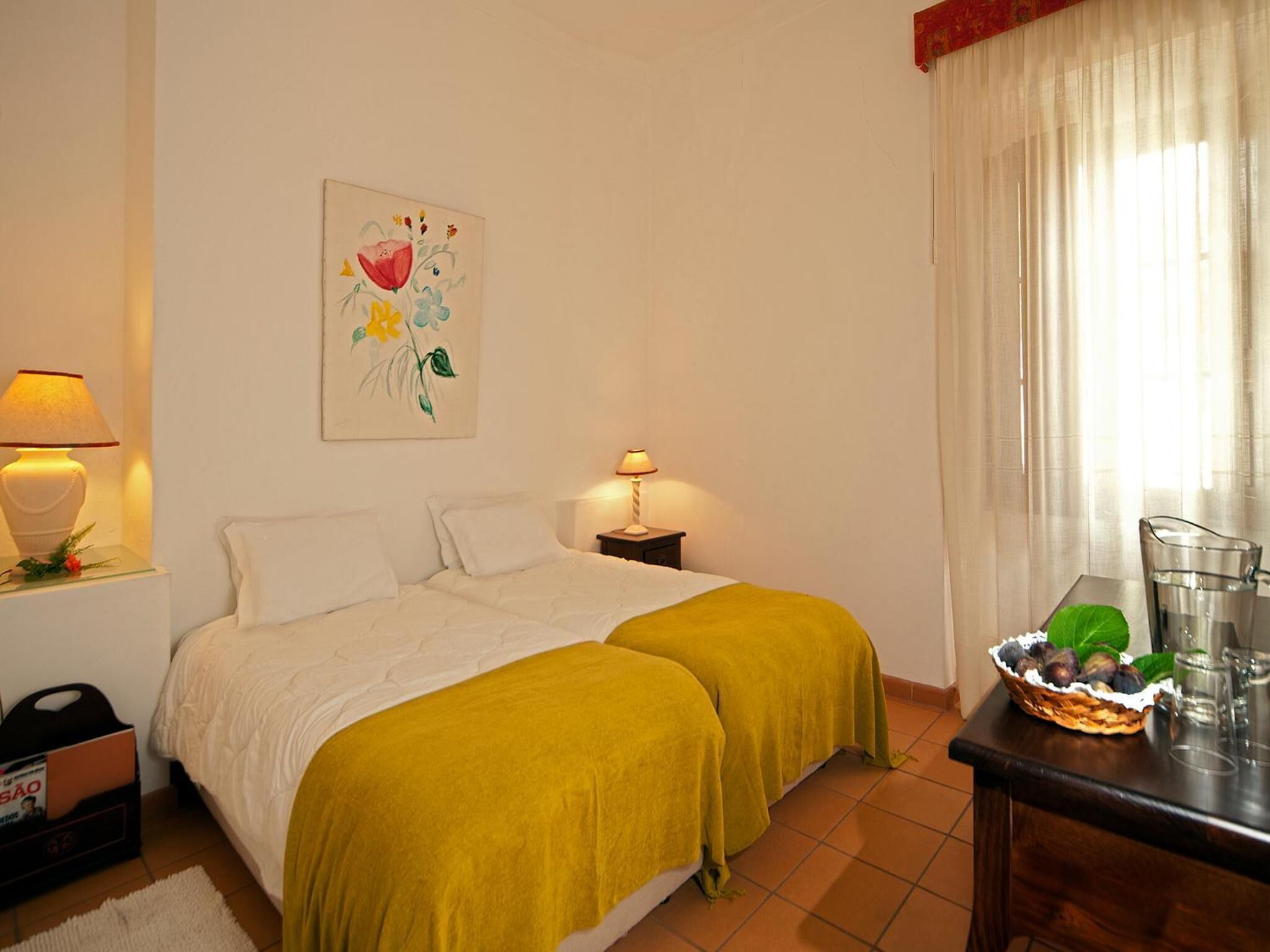 Habitación de hotel con una cama, dos lámparas de noche, un cuadro en la pared, una ventana con cortinas, una mesita con una cesta de fruta y una mesita de noche con lámpara.