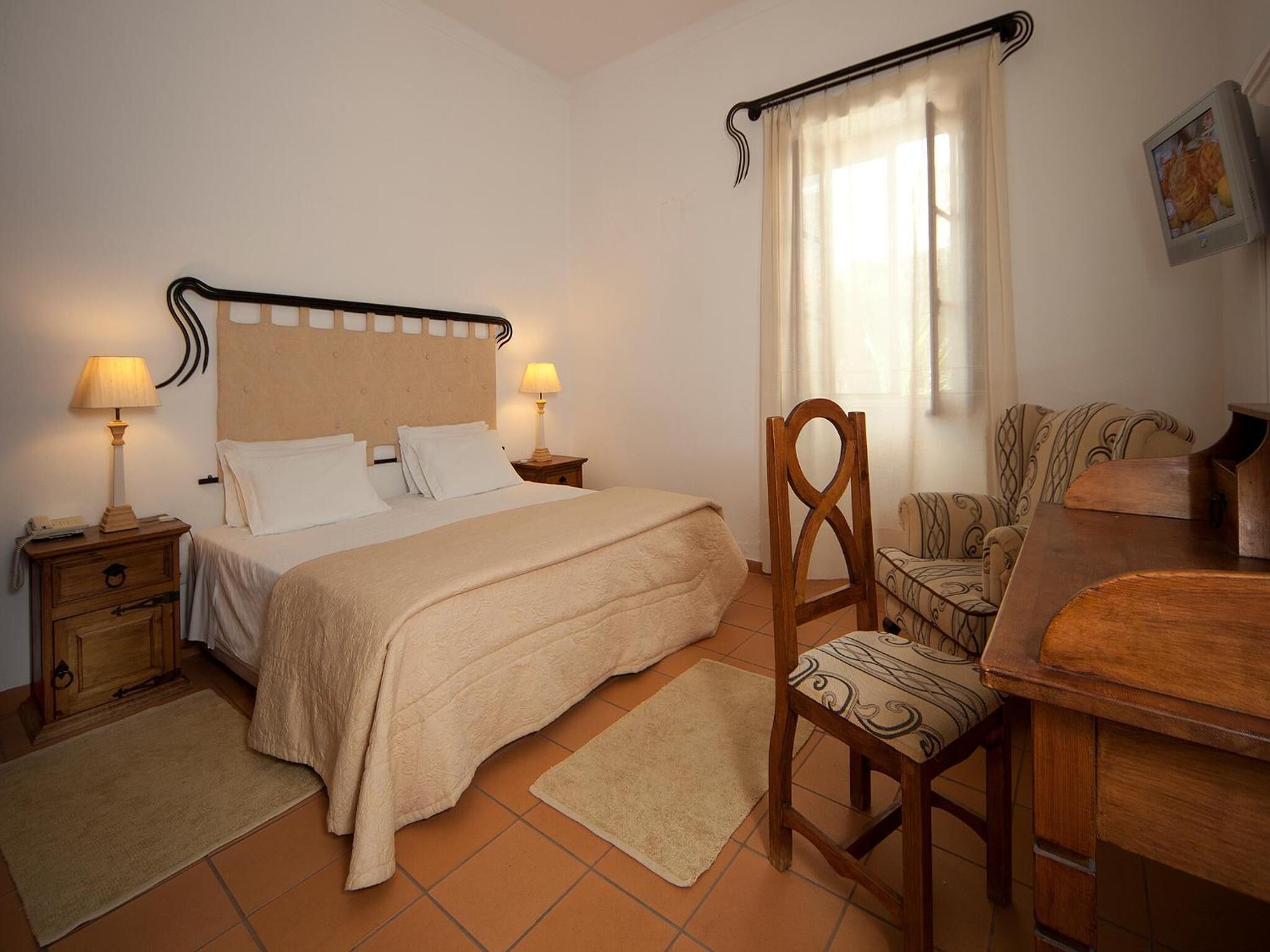A bedroom with a bed, two bedside tables with lamps, a wooden chair, a wooden desk, a patterned sofa, and a window with curtains.