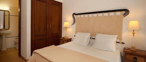 Habitación de hotel con una cama grande, dos mesitas de noche con lámparas, un armario, un baño con lavamanos y espejo, y una lámpara en el techo.